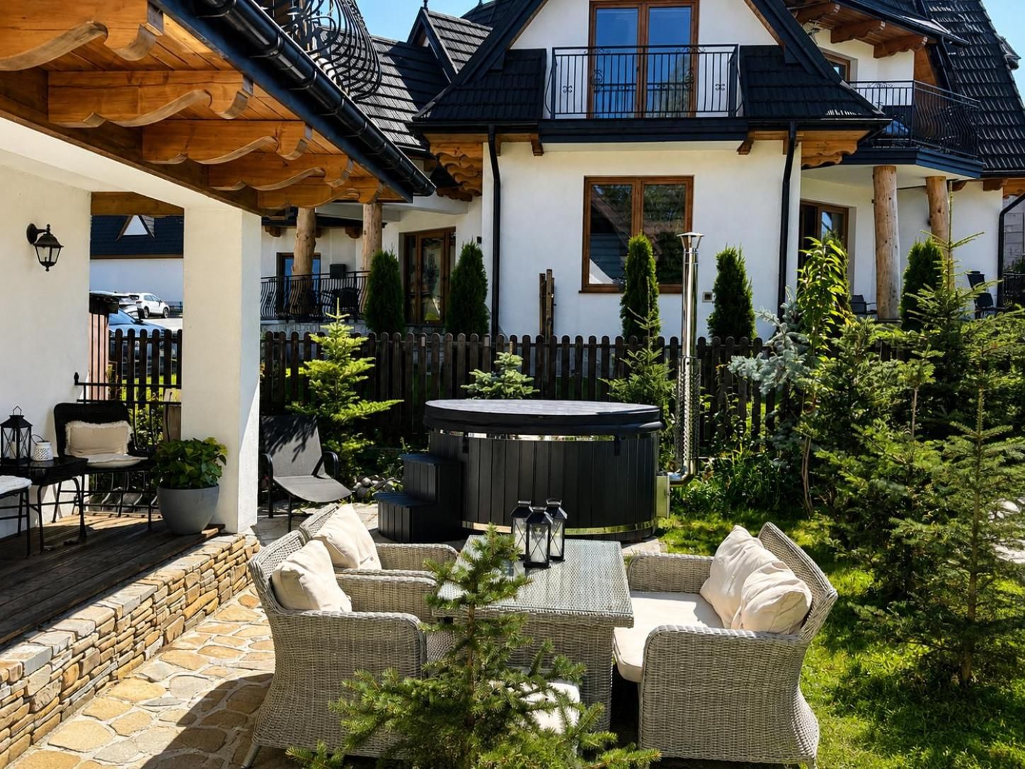 Ogród Czernik House w słońcu: wiklinowe fotele, gorąca balia i biały dom z drewnianymi okapami